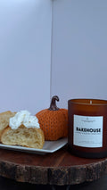 An amber glass jar candle labeled 'BAKEHOUSE' sits on a rustic wood slice table next to a plate with a pastry and a small orange pumpkin against a plain light background.