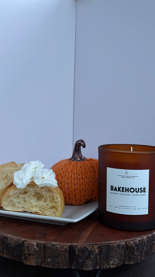 An amber glass jar candle labeled 'BAKEHOUSE' sits on a rustic wood slice table next to a plate with a pastry and a small orange pumpkin against a plain light background.