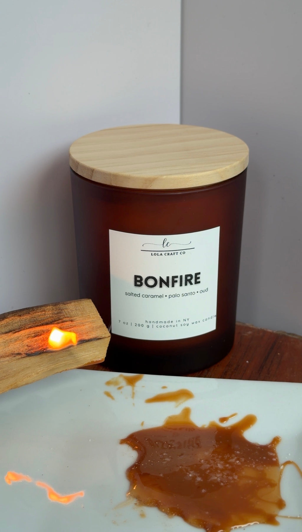 A large amber glass candle jar with a light wood lid and a white label reading BONFIRE, set on a wooden surface next to a lit candle flame. A white plate with caramel-like residue is in the foreground against a plain wall.