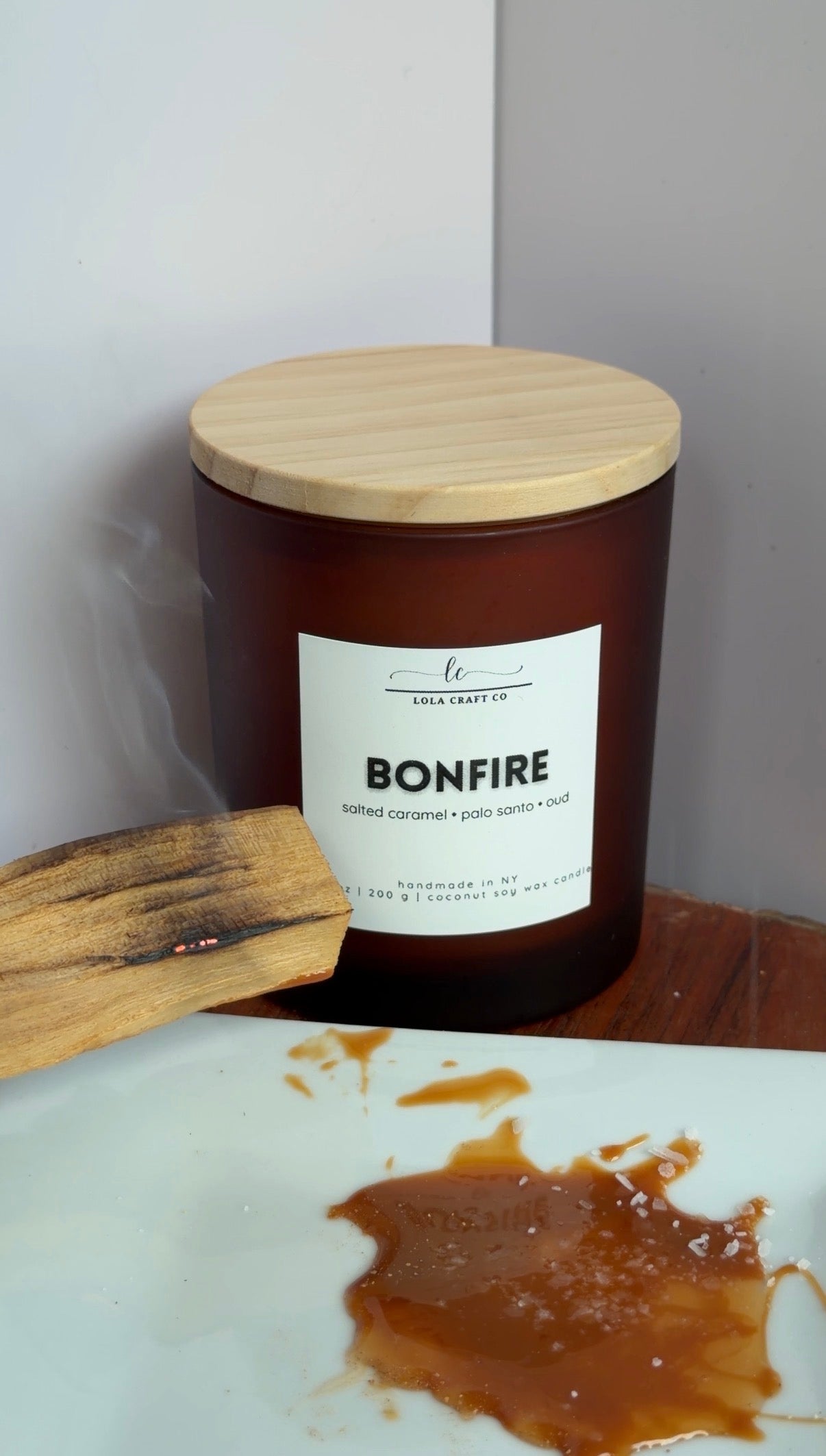 An amber glass candle jar with a light wood lid and a white label reading BONFIRE, set on a wooden surface next to a charred wooden paddle on a white plate with caramel-like residue. Visible brand text on the label reads 'Lea Craft Co'.