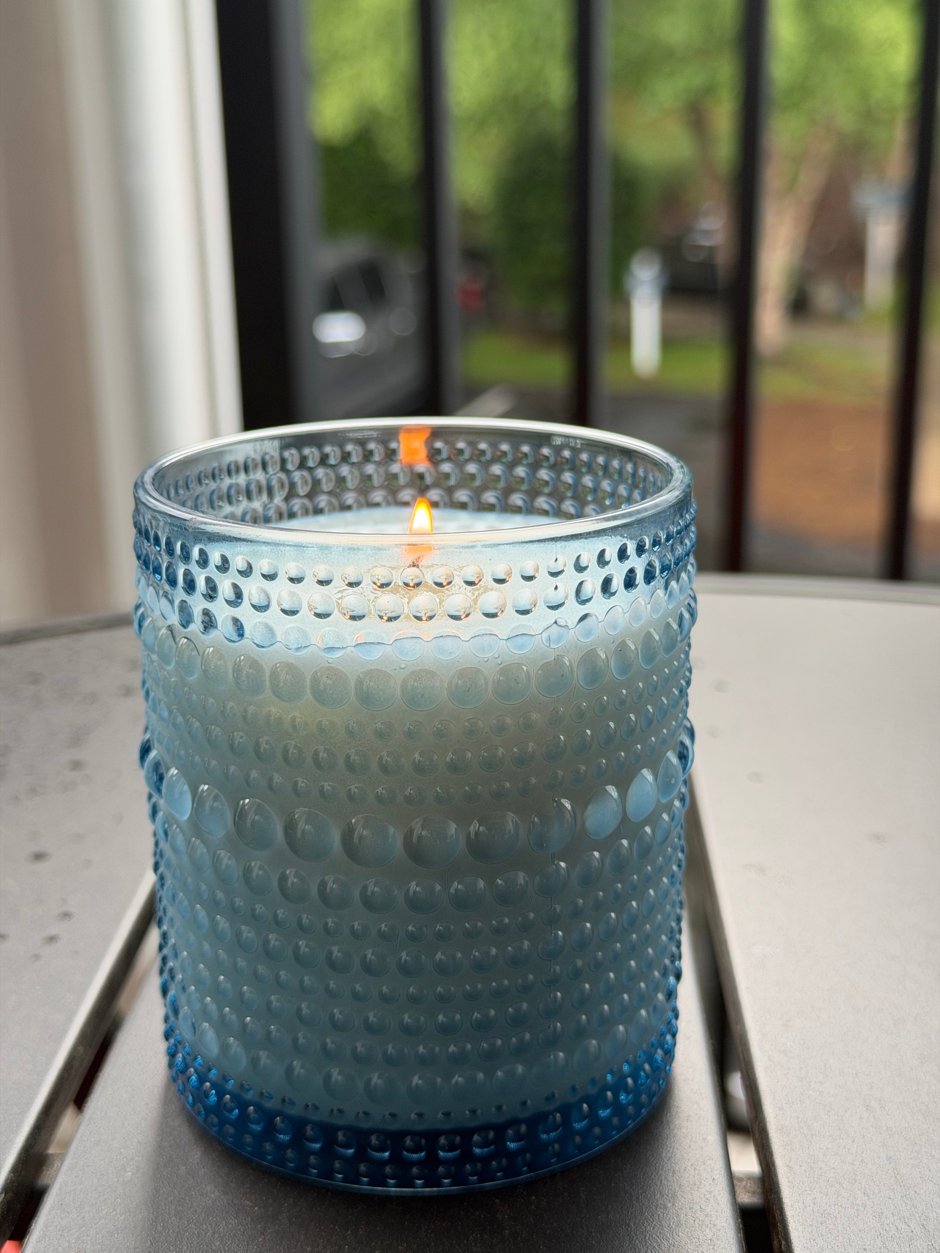 A lit blue textured glass candle holder with a small orange wick, sitting on a gray metal table in front of a window with a blurred outdoor background.