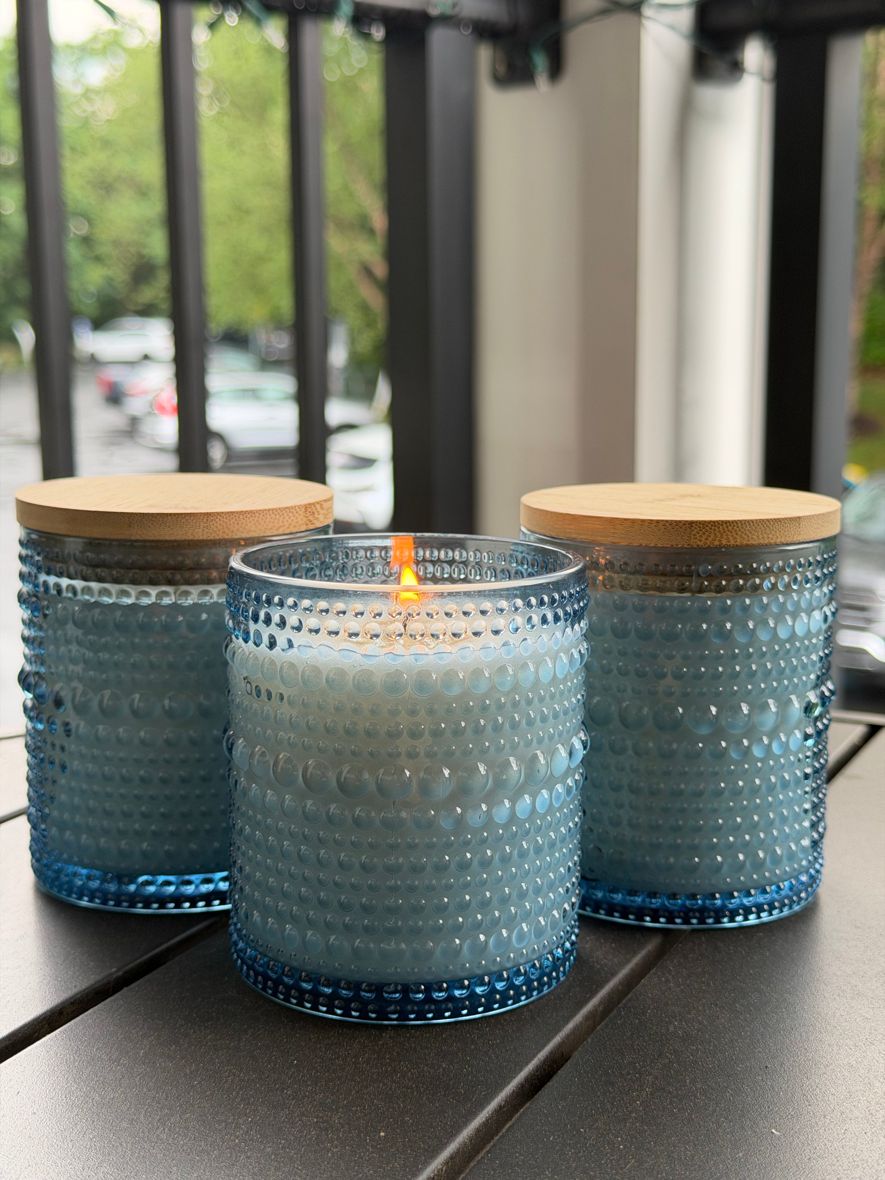 Three blue textured glass candle jars with bamboo lids, with the center candle lit, displayed on a dark outdoor table.