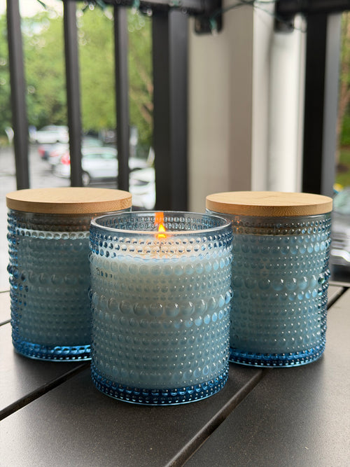 Three blue textured glass candle jars with bamboo lids, with the center candle lit, displayed on a dark outdoor table.