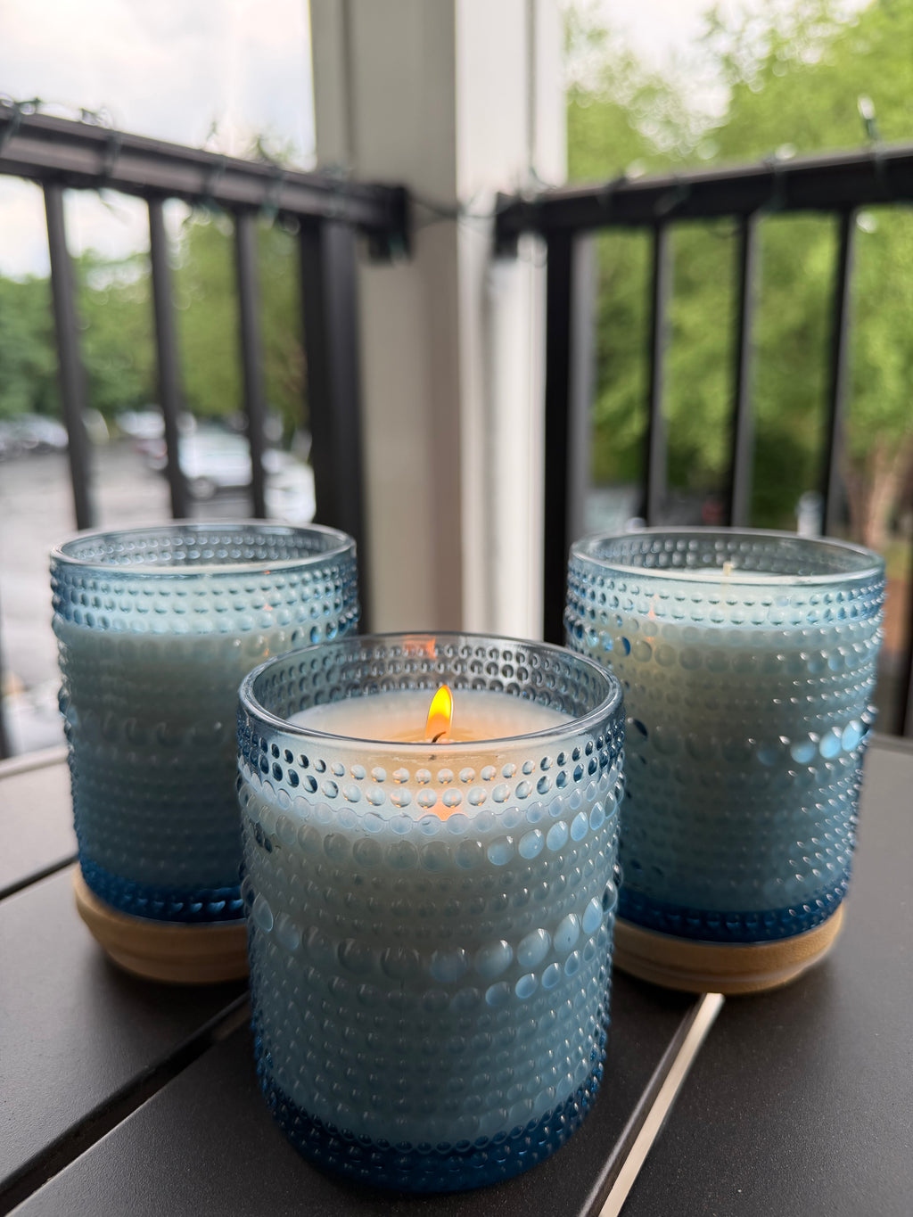 Three blue textured glass candle holders on a dark table, with one holder in the front holding a lit candle and two more holders on either side. A blurred outdoor balcony railing and greenery are visible in the background.