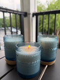 Three blue textured glass candle holders on a dark table, with one holder in the front holding a lit candle and two more holders on either side. A blurred outdoor balcony railing and greenery are visible in the background.