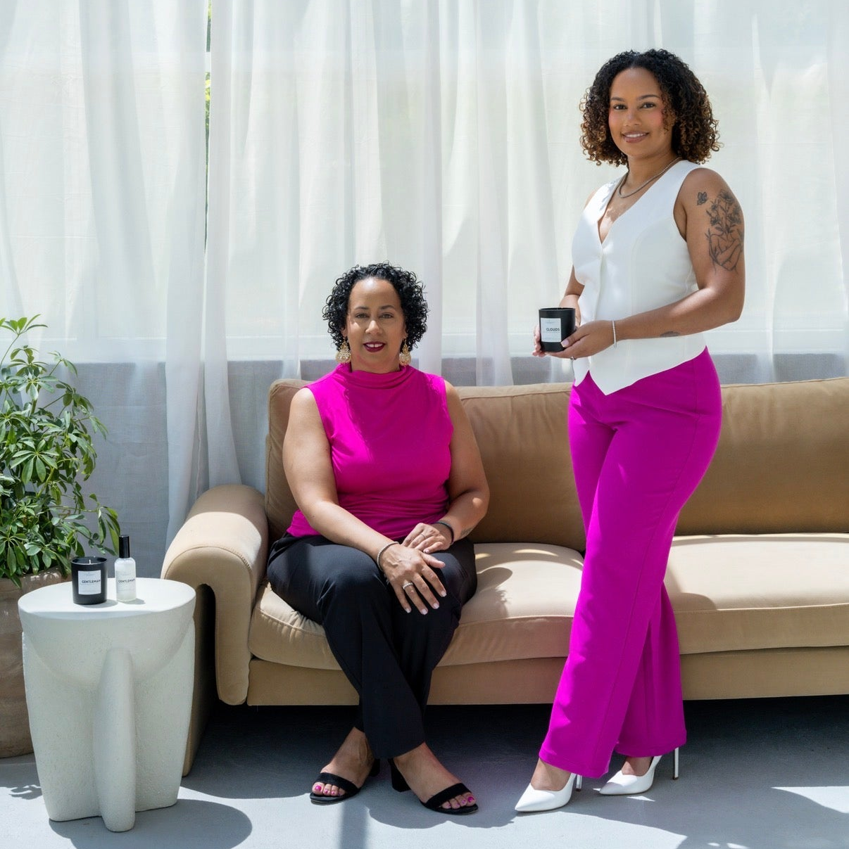 Two women in a bright, modern room with sheer white curtains and a beige sofa. One woman sits on the sofa wearing a bright pink sleeveless top and black pants, while the other stands wearing a white sleeveless top and matching bright pink pants.
