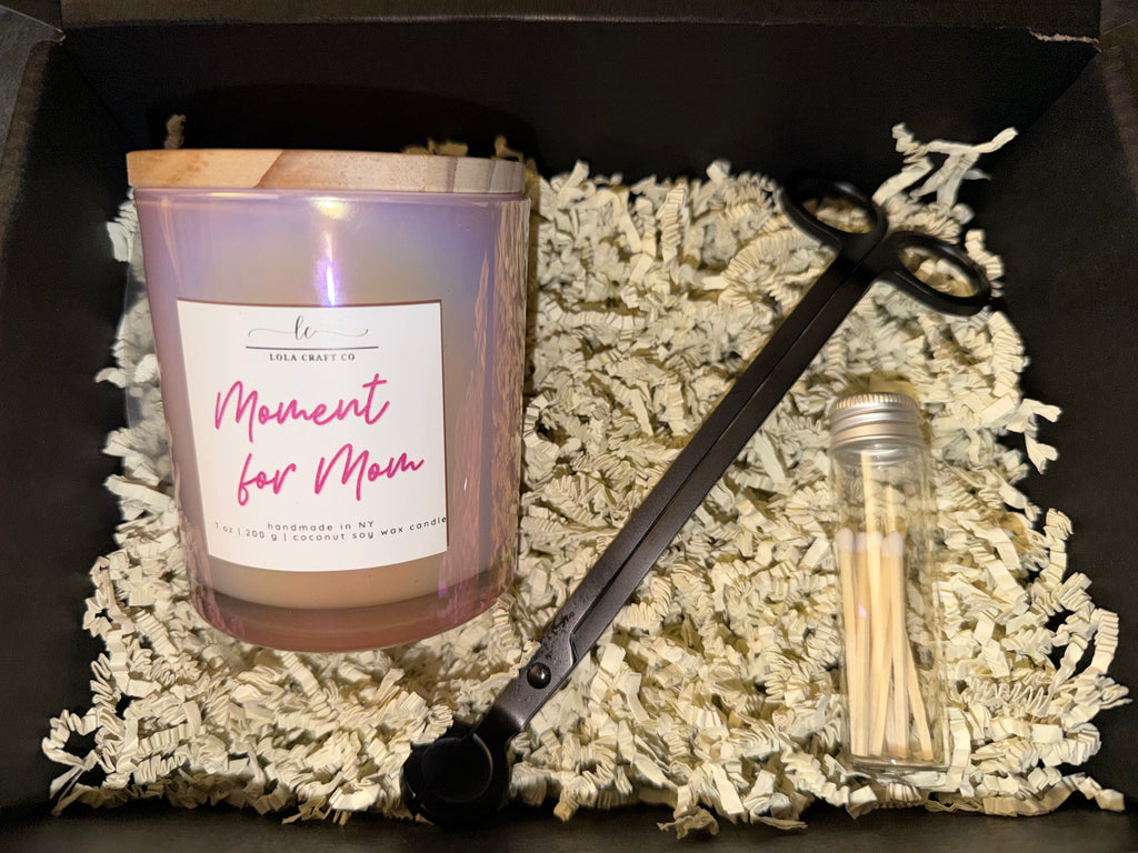 Candle with 'Moment for Mom' label, wick trimmers, and matches on a textured surface