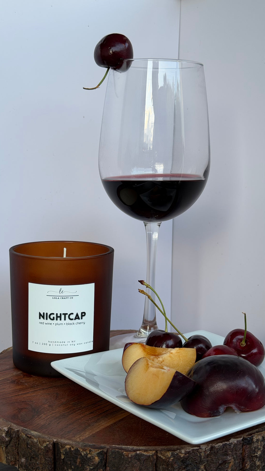 An amber glass candle jar labeled NIGHTCAP sits on a wooden surface beside a white plate with sliced peach, plums, and cherries, with a stemmed wine glass filled with red wine and a cherry garnish in the background.