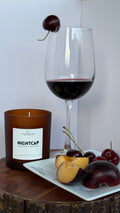 An amber glass candle jar labeled NIGHTCAP sits on a wooden surface beside a white plate with sliced peach, plums, and cherries, with a stemmed wine glass filled with red wine and a cherry garnish in the background.