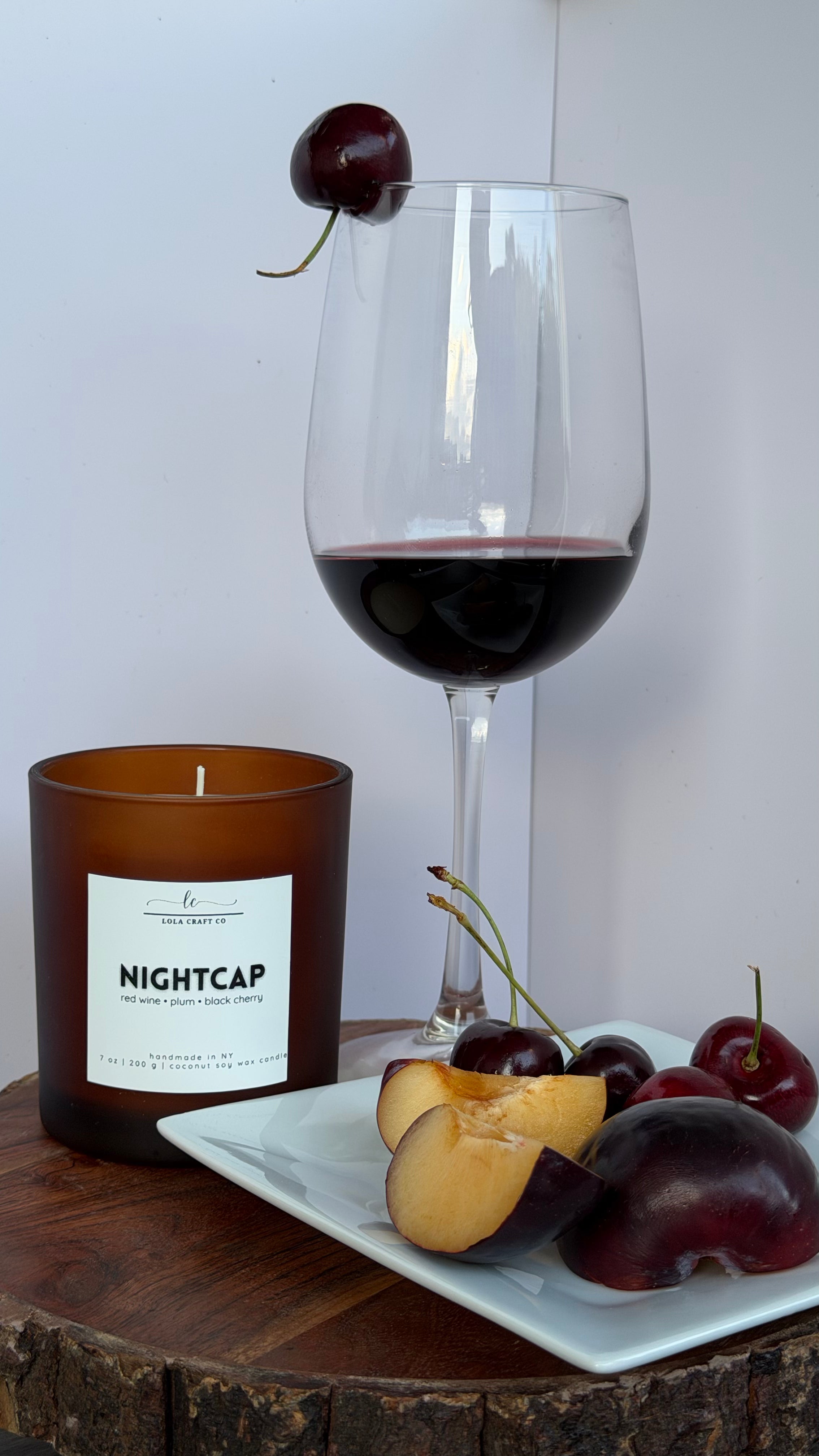 An amber glass candle jar labeled NIGHTCAP sits on a wooden surface beside a white plate with sliced peach, plums, and cherries, with a stemmed wine glass filled with red wine and a cherry garnish in the background.