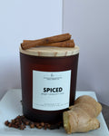 A dark amber glass jar candle with a light wood lid, labeled 'Spiced' by Lola Craft Co, styled with cinnamon sticks, cloves, and fresh ginger on a white surface against a neutral background.