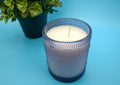 Candle in a textured glass holder on a blue background with a plant.