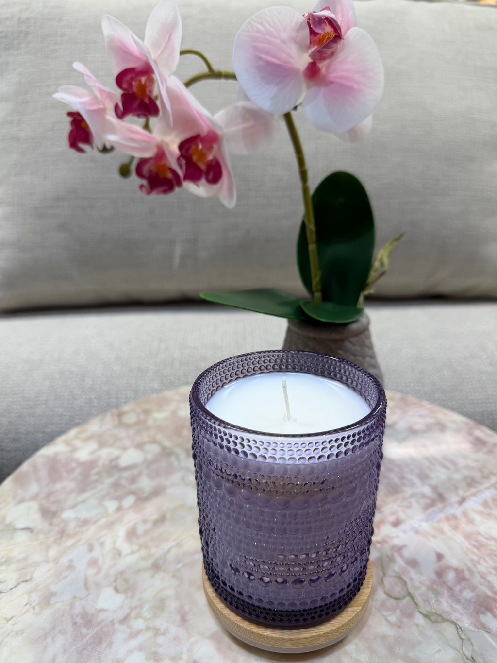 Candle in a textured purple holder with pink orchids on a marble surface.