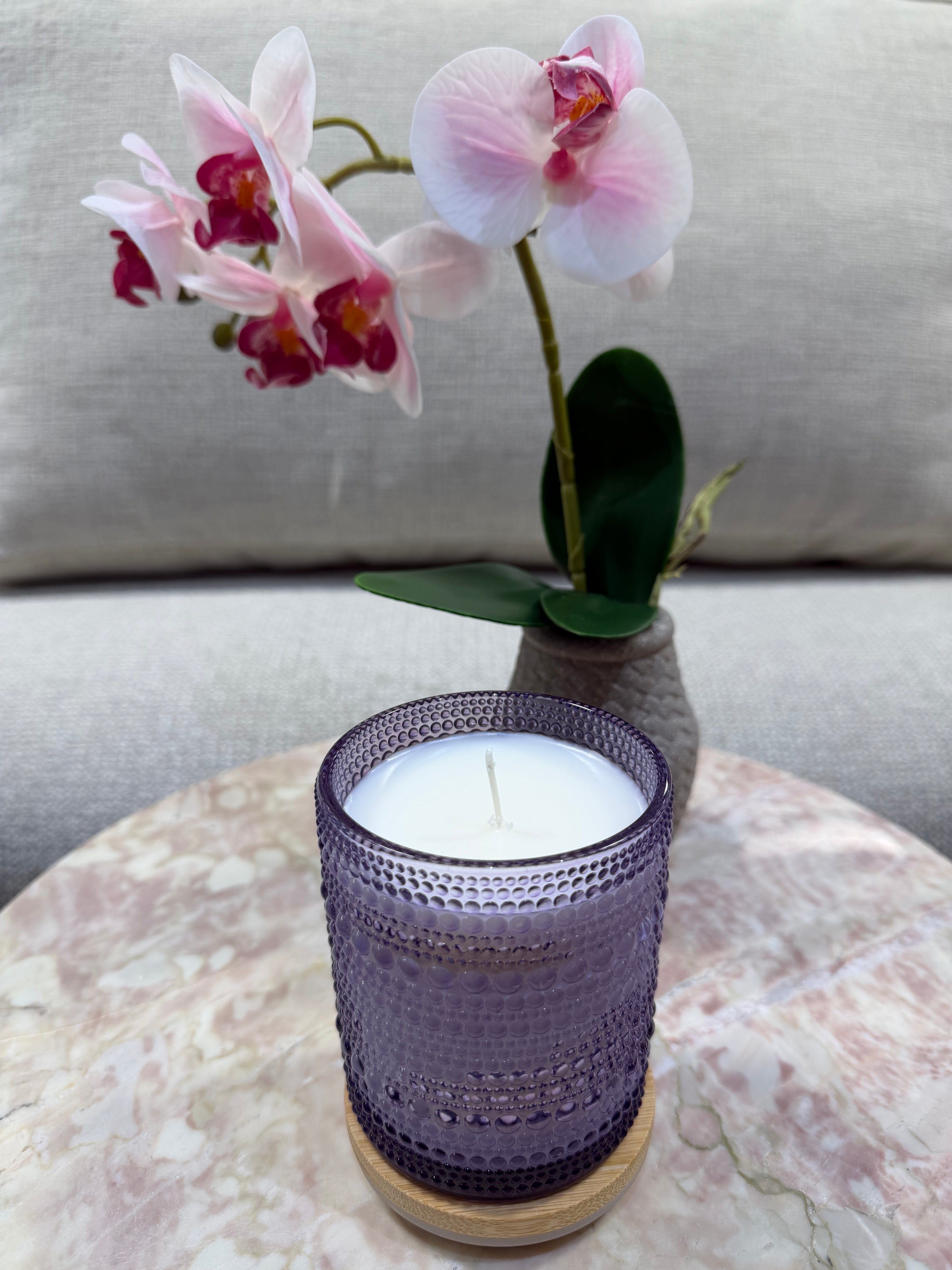 Purple candle in a textured glass holder with pink orchids on a marble surface.