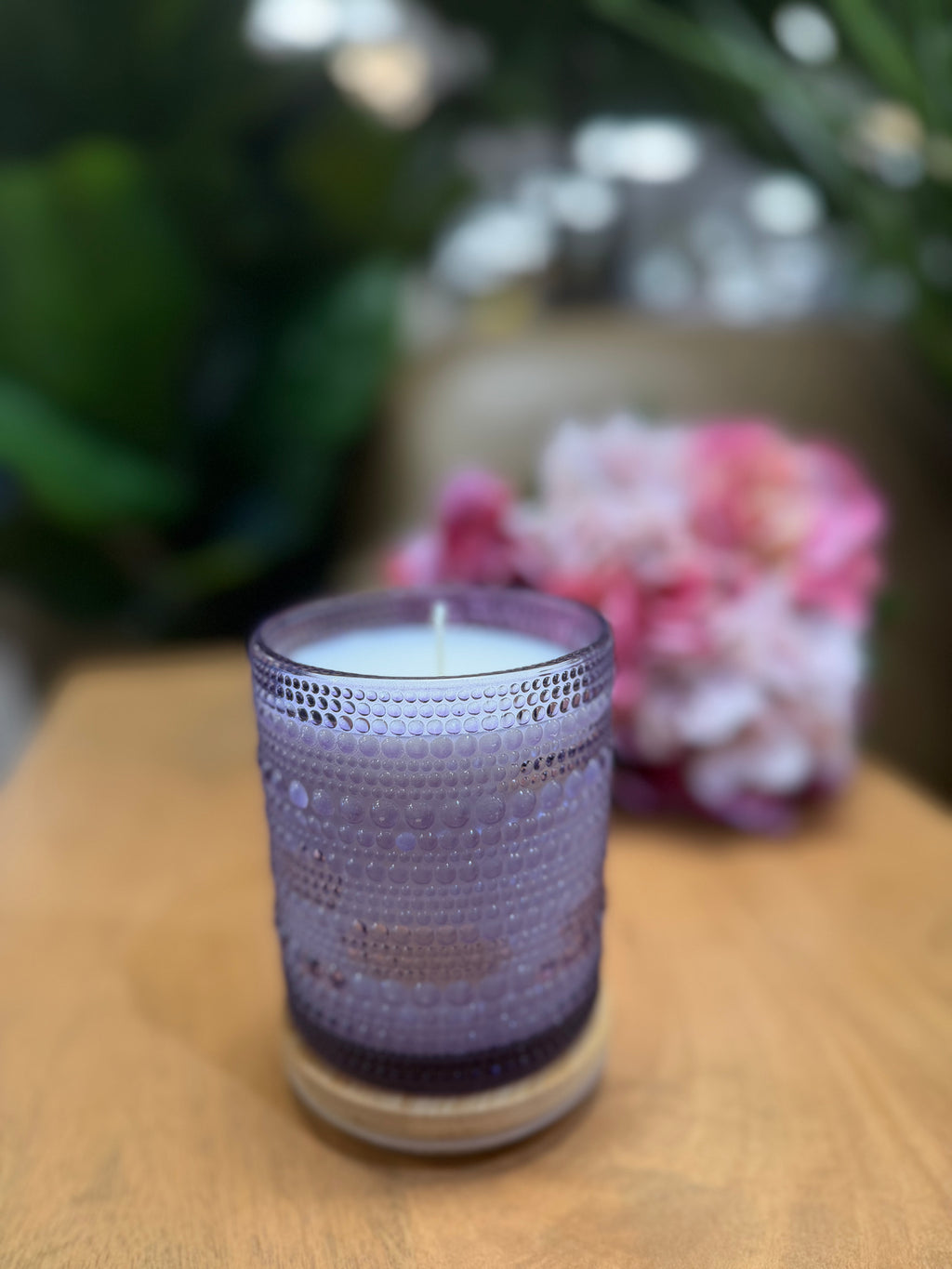 Purple candle in a textured glass holder on a wooden surface with blurred flowers in the background