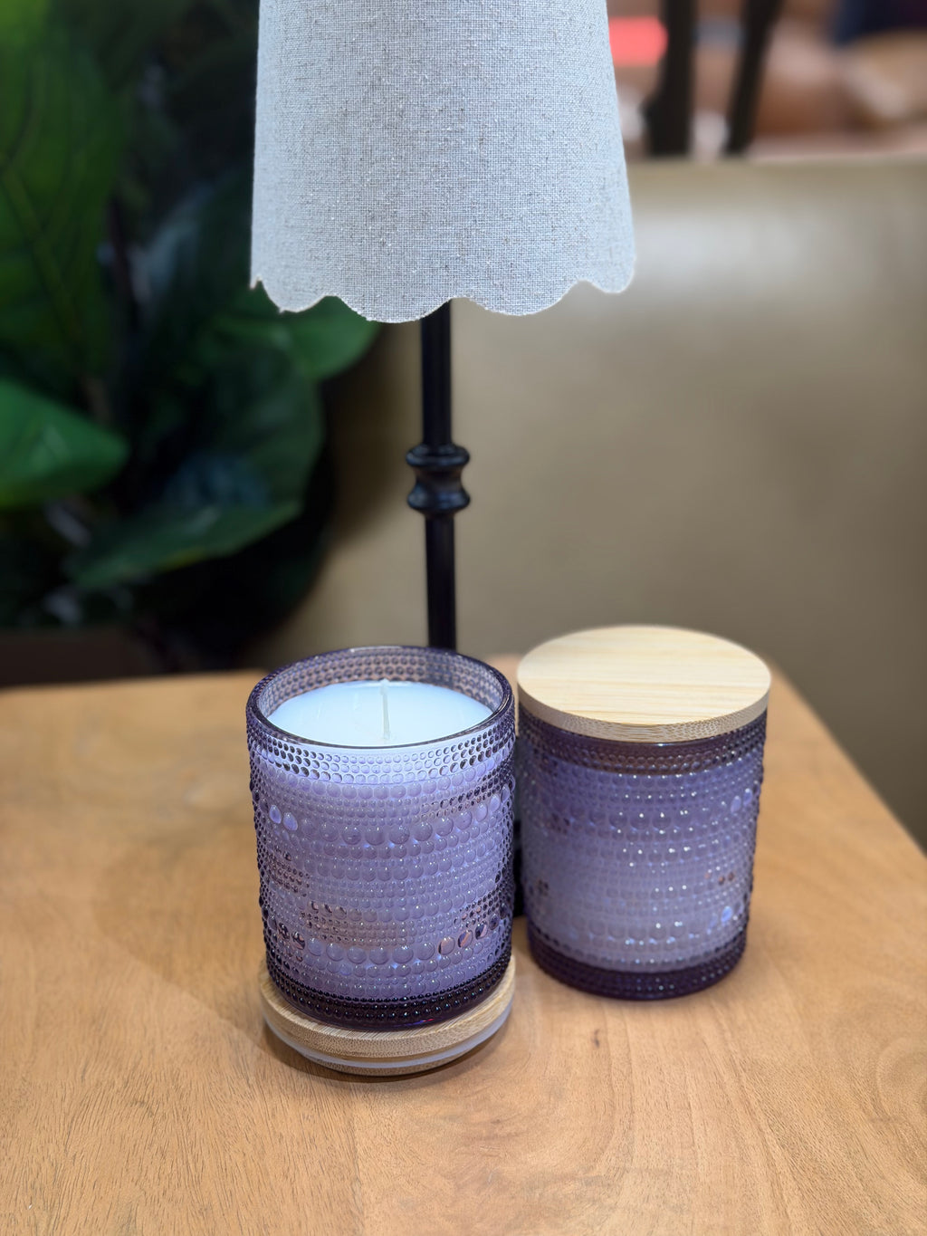 Two purple textured candles on a wooden surface with a lamp in the background.