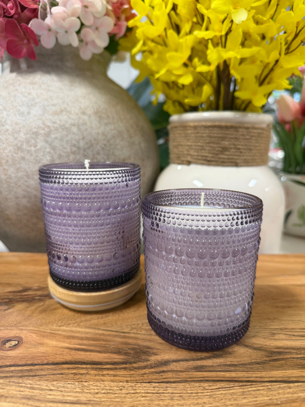 Two purple textured candles on a wooden surface with floral arrangements in the background.