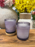 Two purple textured candles on a wooden surface with floral arrangements in the background.