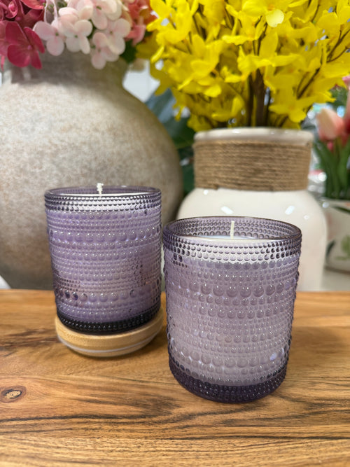 Two purple textured candles on a wooden surface with floral arrangements in the background.
